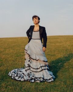 For Styles, his friend and muse Gucci’s Alessandro Michele crafted a frothy, lace-trimmed creation, paired here with a double-breasted tuxedo jacket. Styles is seen standing on an empty green hill.